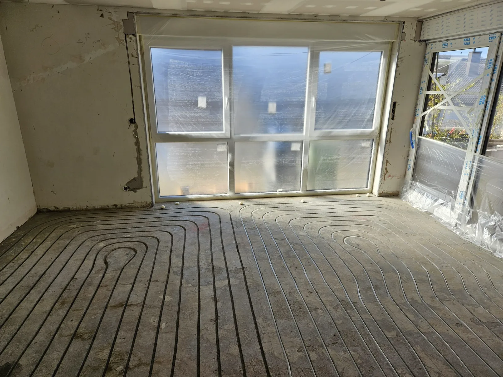 Wohnraum im Rohbau mit Fensterfront: die Heizrohre liegen in schneckenförmig gefrästen Kanälen im Estrich.